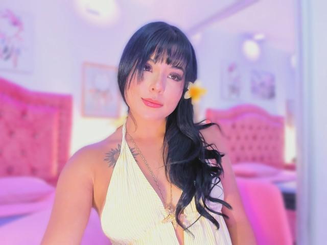 GraceClarke with black hair, pink lips, and a sunflower in her hair, wearing a yellow halter top, showing her tits and a tattoo on her chest. Bedroom background.