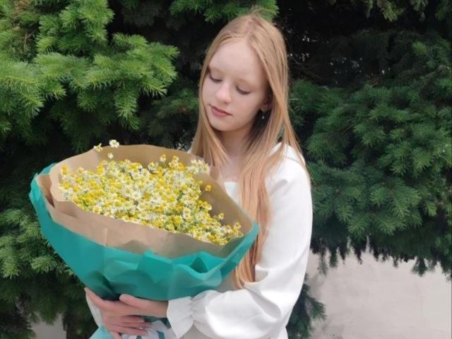 Tara Capistrán, con cabello rubio largo, sosteniendo un ramo de flores amarillas envuelto en papel verde, vestida con una camisa blanca de manga larga, mirando hacia abajo con una expresión suave..