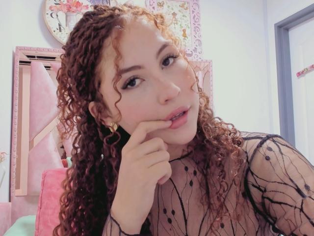 IrisDalton with curly red hair, pale skin, and black see-through top, biting her finger, showing off her tits, wearing gold earrings, looking cute and sexy in a selfie.