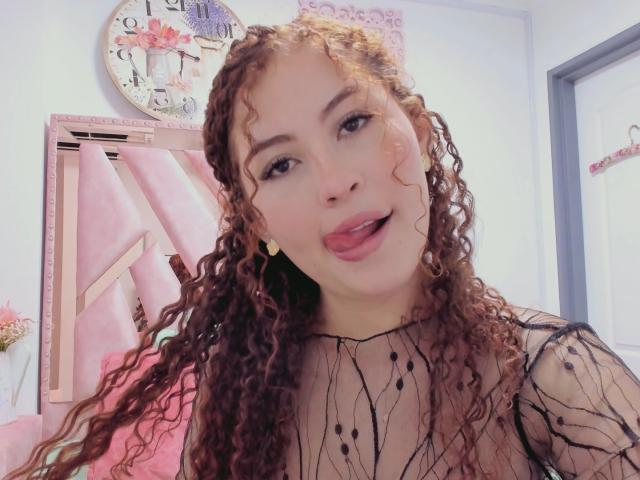 IrisDalton with curly brown hair, wearing a sheer black top, pouty pink lips, and yellow earrings, sticking her tongue out. She's cute and has a playful expression.