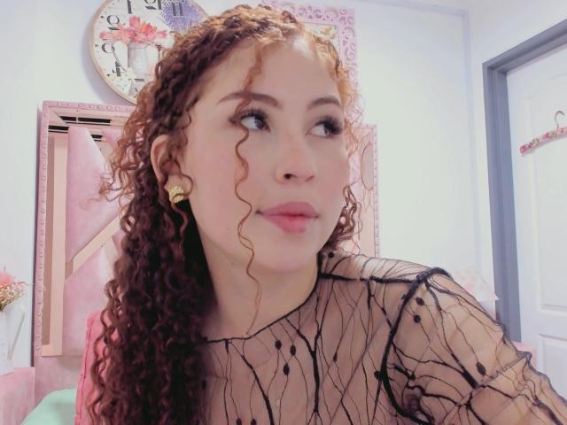 IrisDalton with curly brown hair, wearing a sheer black top with black lines, has a cute face, small nose, and pink lips, standing in a pink room.