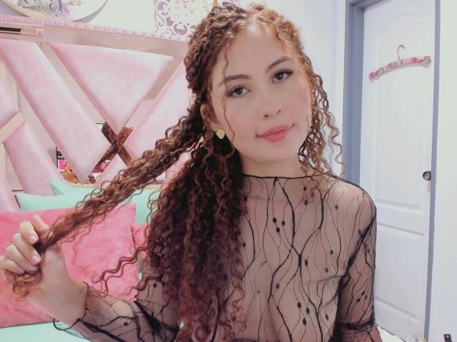IrisDalton with long curly brown hair, wearing a sheer black mesh top, showing small tits, pink lips, light skin, and gold earrings, holding her hair, smiling.