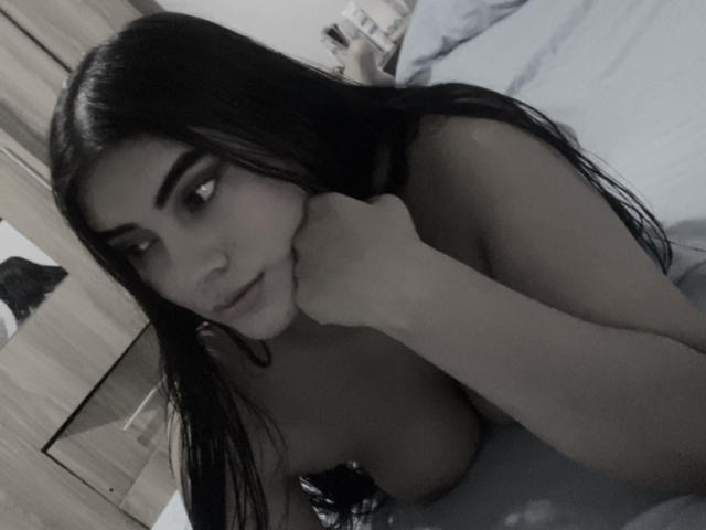 AlaraKey with long black hair, looking at camera, resting chin on hand, topless, showing large boobs, soft light, bedroom, grayscale, slightly blurry, subtle smile.