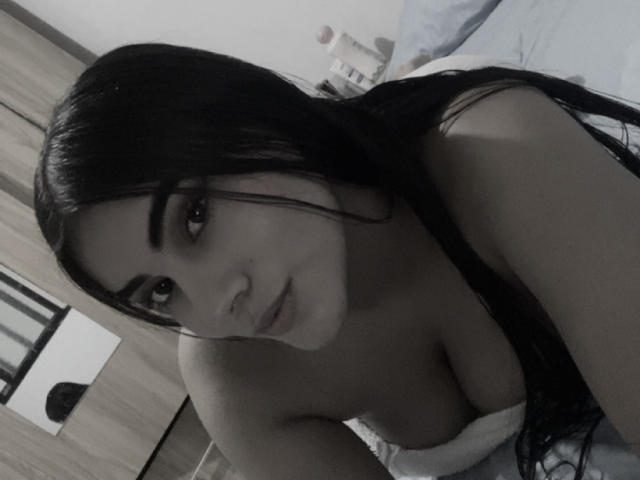 AlaraKey with long black hair, brown eyes, and large tits, looking at the camera with a slight smile, lying on a bed, in a grayscale selfie.