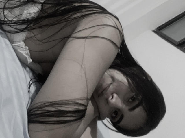 AlaraKey with long dark hair, topless, lying on bed, head tilted, arm over chest, looking at camera, soft black-and-white selfie, showing small breasts, slight smile.