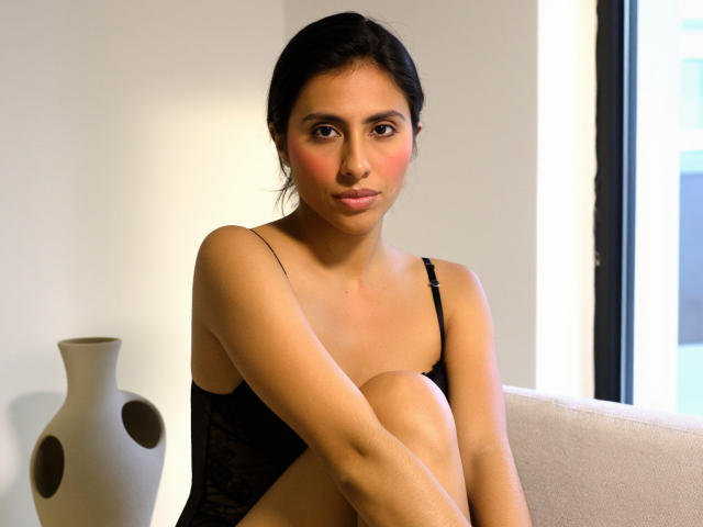 JeanieWise, com cabelo escuro, pele bronzeada e blush rosa nas bochechas, vestindo um sutiã de renda preto, sentada com os joelhos dobrados, com um vaso branco com furos à esquerda..