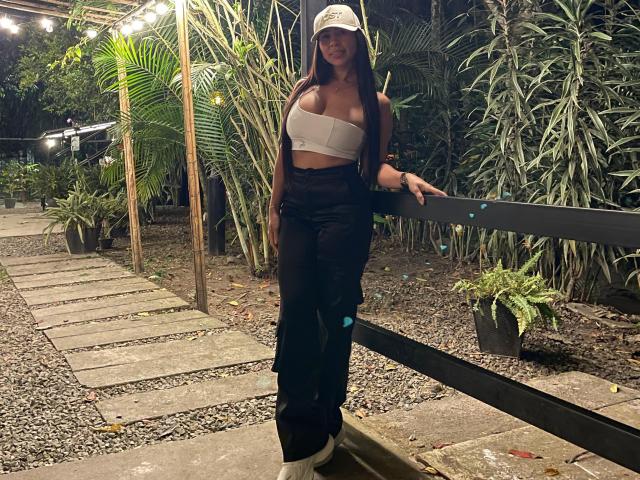 NahomyStone with large tits, wearing a white strapless top, black pants, and a cap, leaning on a black bench in a nighttime garden with tall green plants.