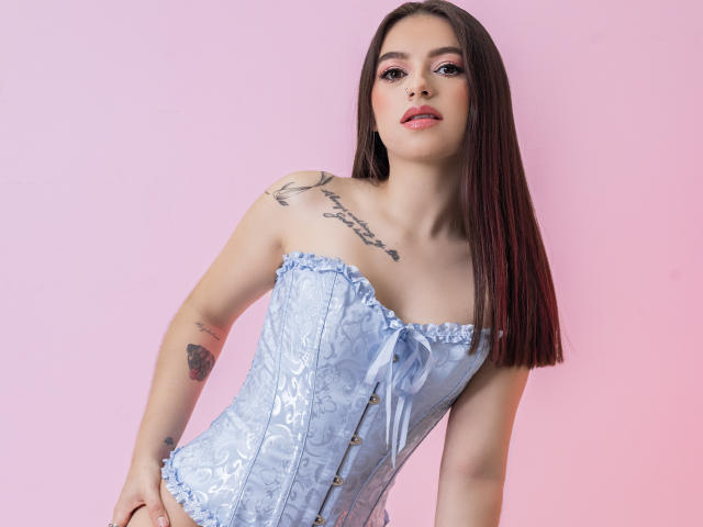 PaolaYKatty with long brown hair, wearing a light blue lace-up corset, showing small tits, tattoos on her shoulder and arm, standing against a pink background.