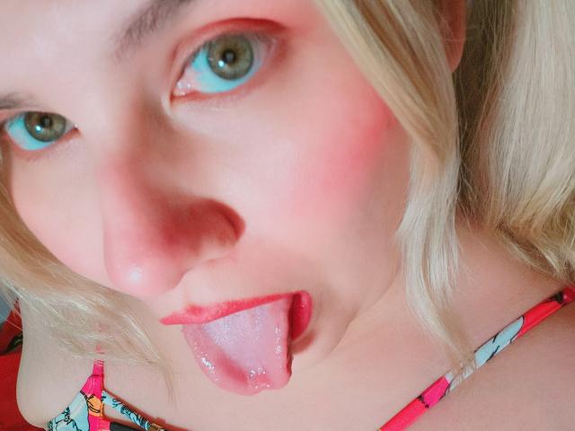 Akkanee's face, blonde pigtails, blue eyes, red lipstick, and tongue out. She's sticking out her wet, shiny pink tongue. She's wearing a colorful, patterned top.