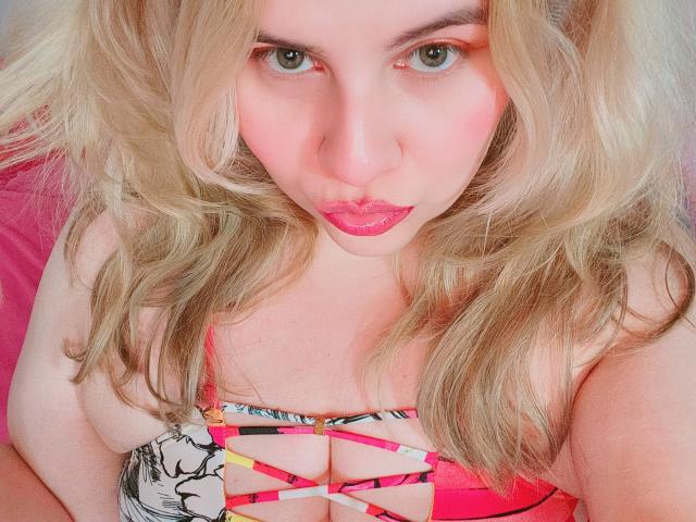 Akkanee with blonde wavy hair, pink lipstick, and green eyes, looking at the camera, wearing a pink and black strappy top, showcasing her large, shiny tits.