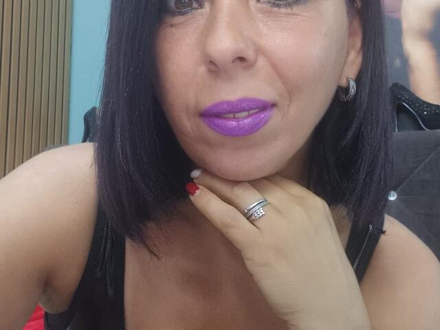 LauraSin with black hair, purple lipstick, red nail polish, black tank top, silver ring, and earrings, holding her chin, showing her ass.