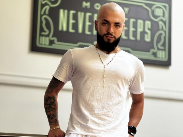OscarWills, bald with a thick black beard, white t-shirt, gold chain, tattooed arm, black watch, standing, looking at camera, white wall, green sign in background.