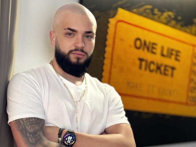 OscarWills, mit einem dicken Bart, trägt ein weißes T-Shirt, eine goldene Kette und eine Armbanduhr, die Arme sind verschränkt, er hat ein Tattoo am rechten Arm, einen Nasenring und einen ernsten Gesichtsausdruck..