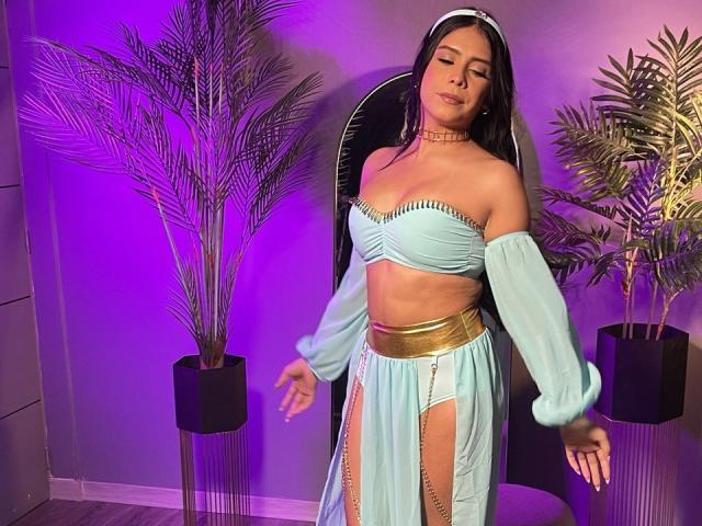Mademoiselle. Con una blusa azul claro de corte asimétrico, un cinturón dorado y una falda fluida, lleva un collar y una diadema, y está de pie frente a plantas en macetas con iluminación púrpura..