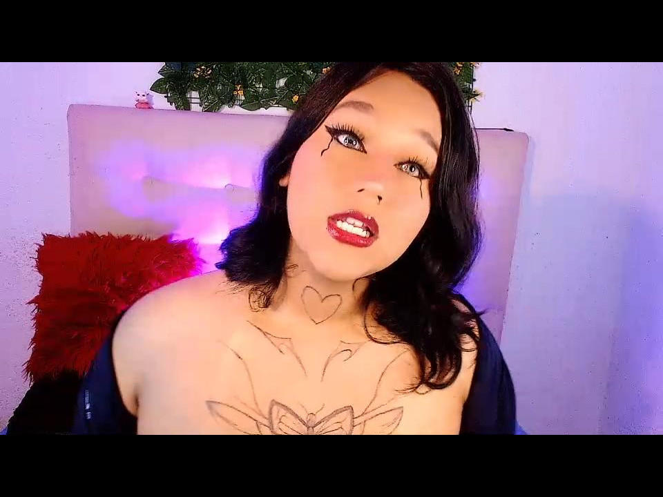 SalomeMirandaa with black hair, red lipstick, and tattoos on her chest and neck, wearing a black top, looking up with parted red lips. Purple and pink lights in background.