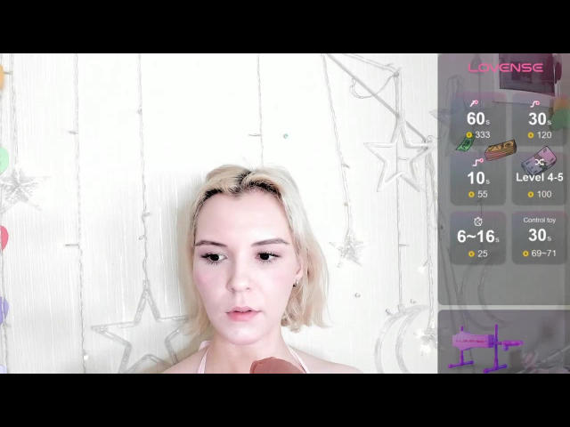 KayaDevis has short blonde hair, pale skin, and a neutral expression. She's looking slightly to the side with a camera in front of her. Her tits are small and she's topless.