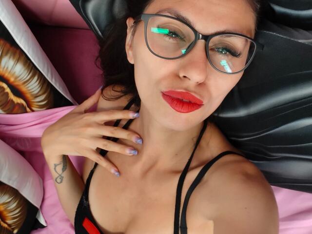 DanaWaw with red lipstick, black glasses, and painted nails, lying on a black pillow, showing her tits in a black strappy top, hand on her neck, smiling.