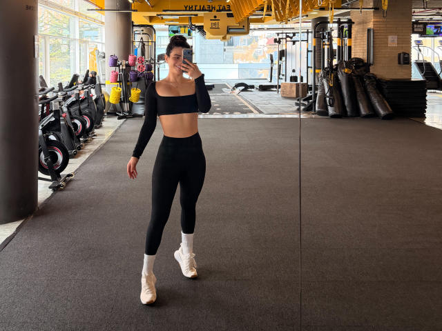 PaulinaValley in a gym, wearing a black crop top and high-waisted black leggings, white socks, and white sneakers, taking a mirror selfie, showing her slim, fit, small-ass body.