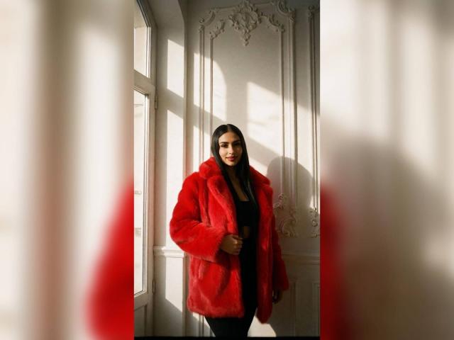 MiilaWx with long dark hair, wearing a red fur coat, black top, and black pants, standing in a sunlit room, casting a shadow on the white wall.
