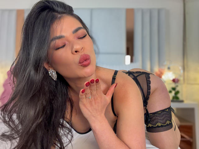 PaulinaValley with long black hair, wearing black lace lingerie, pouting her lips, her red-manicured hand near her face, showing off her earrings and a ring on her finger.