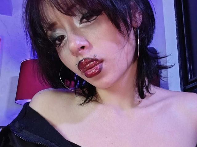 ViolettClay with dark brown hair, hoop earrings, nose piercing, and red lipstick, wearing an off-shoulder black top, looking wistfully to the side.