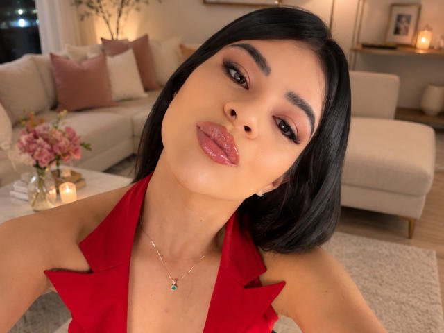 PaulinaValley with glossy pink lips, dark straight hair, and a red halter top, wearing a silver necklace, in a cozy, warmly lit room, looking at the camera.