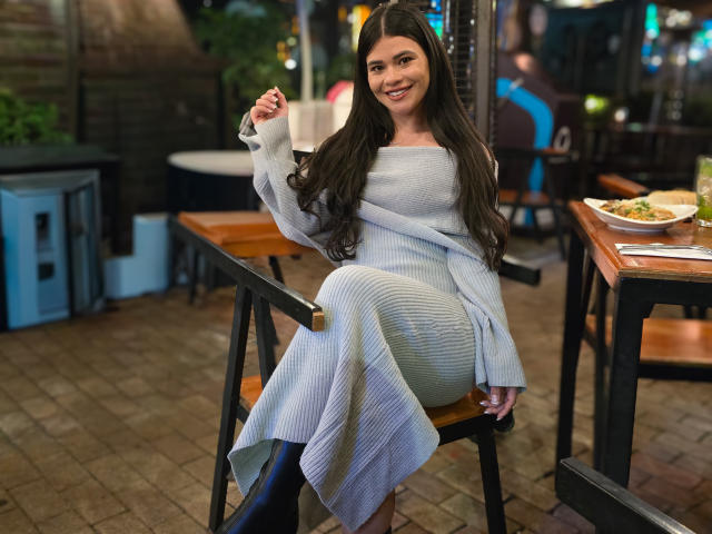 PaulinaValley with long dark hair, smiling, wearing a light gray dress, sitting on a wooden chair, holding a fork, in a dimly lit restaurant with a table in foreground.