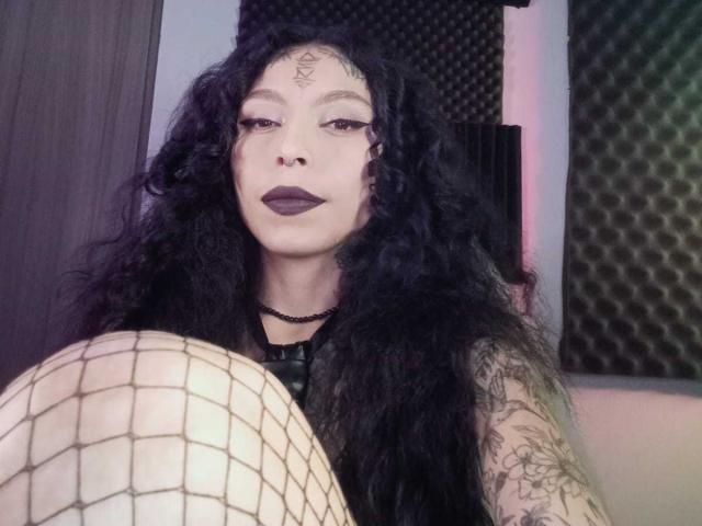 LaurieRzz hat langes, schwarzes, lockiges Haar, dunklen Lippenstift und Netzstrümpfe, die bis zu ihren Knien reichen. Sie trägt eine schwarze Halskette und hat Tattoos an ihrem Arm..