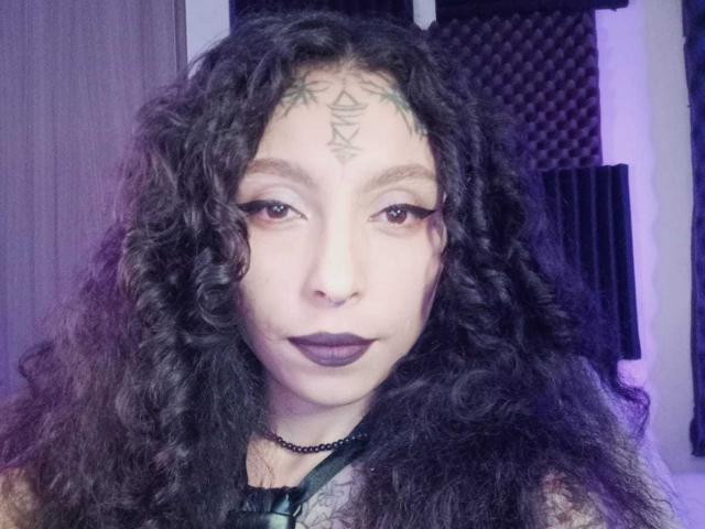 LaurieRzz con capelli neri ricci, tatuaggio a forma di triangolo verde sulla fronte, rossetto scuro, eyeliner spesso, collana nera, che guarda la fotocamera, illuminazione viola-blu..