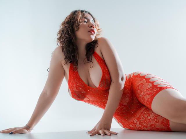 SweetyAliceHotX with curly brown hair, red lace dress, large tits, and cut-out sides, leaning on white floor, eyes closed, red lipstick, and a sexy, confident look.