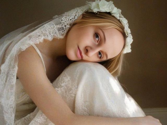 FlyFreedom, with blonde hair and a white lace wedding veil, leans on her knee, looking sad-eyed. She's wearing a lacy white dress and a floral headpiece.