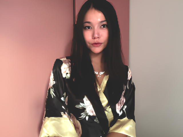 ShelliQuirin with long black hair wearing a black floral kimono with yellow trim, standing against a pink and beige wall, looking at the camera with a subtle smile.