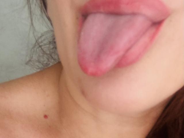 EverlastBlum's tongue is out, pink and wet, with slightly parted red lips. She has a small red mole on her neck. Her skin is light.