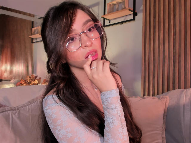 EvaMcAllister with long brown hair, wearing clear glasses and a sheer blue lace top, biting her finger with pink lipstick, sitting on a beige sofa.