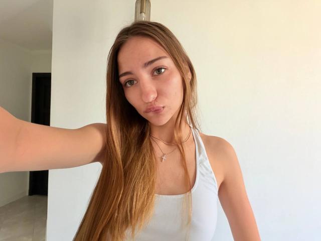 AdrianaNalley with long brown hair, pouty lips, small nose ring, white halter top, necklace, and arm extended for selfie. She's a cute, slender girl with green eyes.