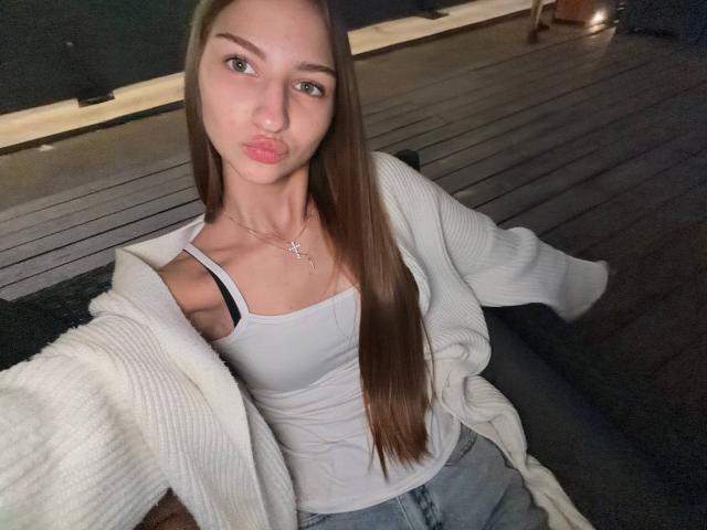 AdrianaNalley with long brown hair, pouty pink lips, wearing a white tank top, black bra strap visible, white cardigan, and blue jeans, sitting on a wooden bench.