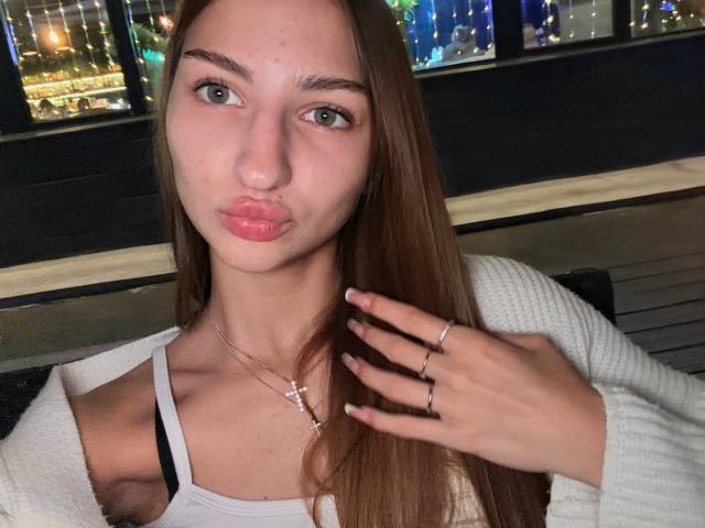 AdrianaNalley with long brown hair, pouty pink lips, green eyes, white tank top, white cardigan, gold necklace, rings, and hand near face, at night.