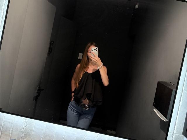 AdrianaNalley with long brown hair, wearing a black off-shoulder top and blue jeans, taking a mirror selfie, showing her small tits and slim ass.