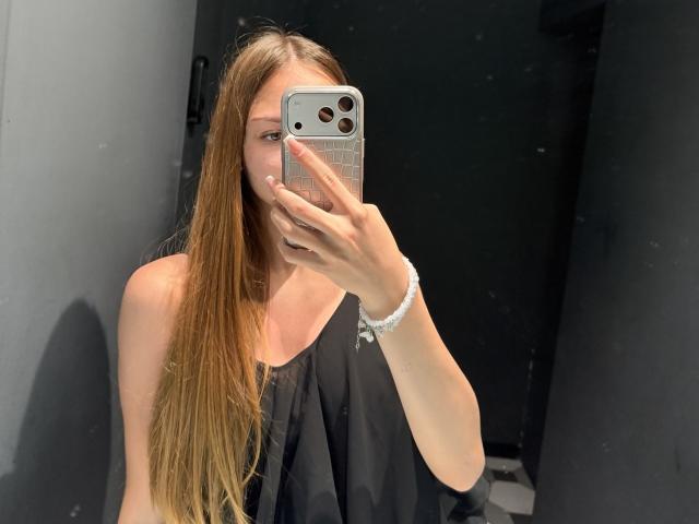 AdrianaNalley with long brown hair, black sleeveless top, holding silver iPhone, taking mirror selfie, white bracelet on right wrist, black door in background.