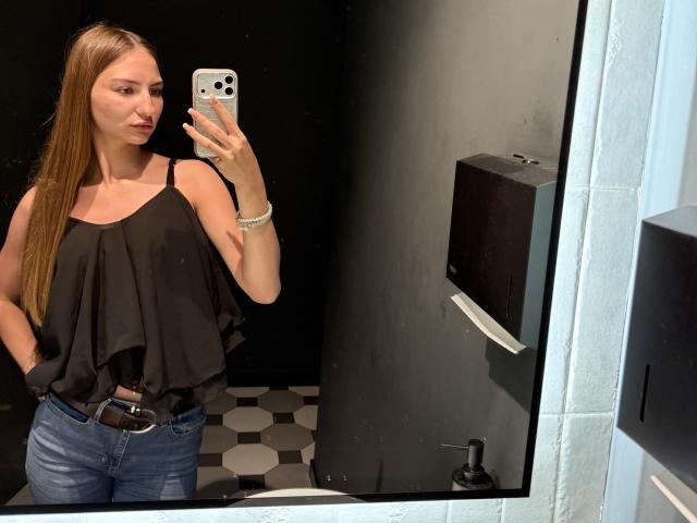 AdrianaNalley with long brown hair, black ruffle top, blue jeans, holding phone in black-walled bathroom, mirror reflection, showing small tits, slim build, serious expression, white bracelet.