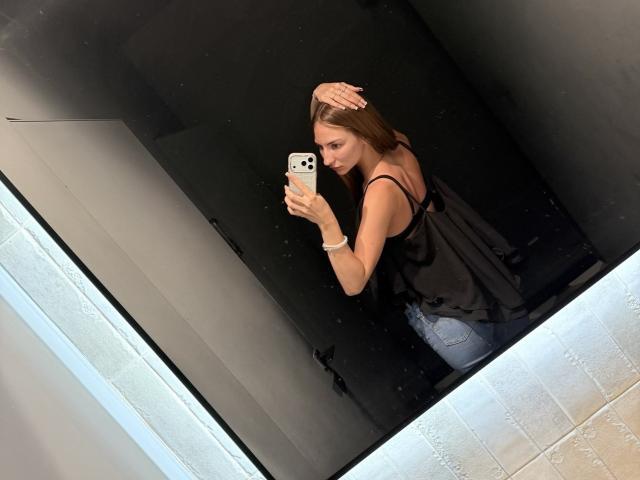 AdrianaNalley with long brown hair, wearing a black sleeveless top and blue jeans, taking a mirror selfie, holding an iPhone, smiling, with her hand on her head.