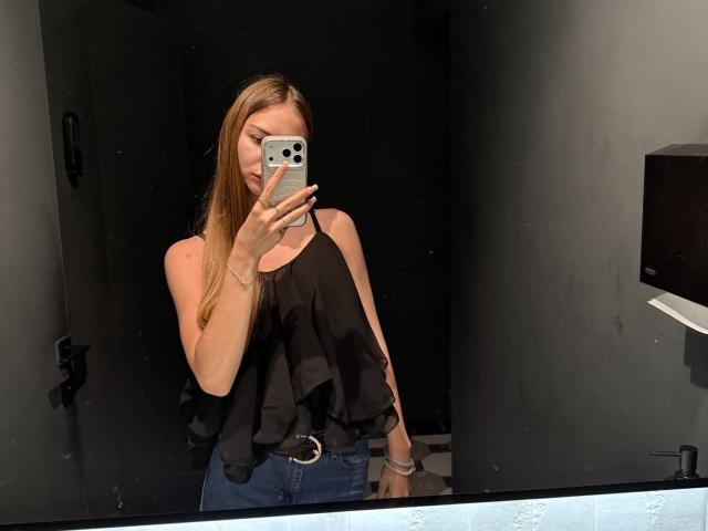 AdrianaNalley with long blonde hair, wearing a black ruffle top and blue jeans, taking a mirror selfie with a silver iPhone, showing her boobs in the reflection.