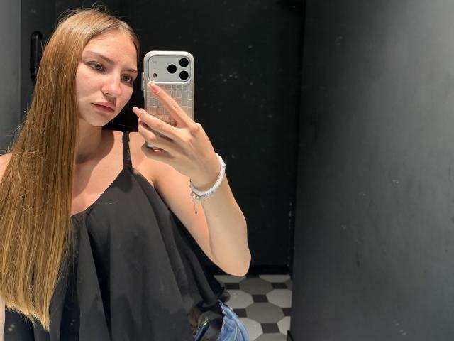 AdrianaNalley with long blonde hair, black off-shoulder top, holding phone, mirror selfie, black walls, white bracelet, hexagon floor tiles, serious expression, phone with white case.