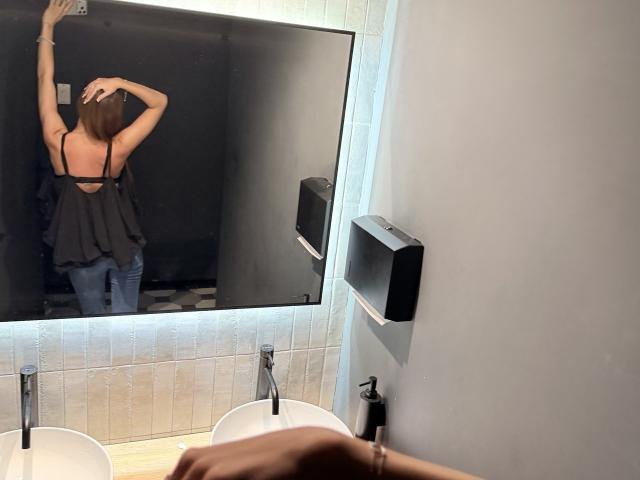 AdrianaNalley's back in a bathroom, wearing a black top with straps and blue jeans, arm raised, reflection in mirror shows her ass and back, hand on sink.