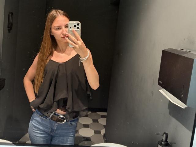 AdrianaNalley with long brown hair, black sleeveless top with ruffles, blue jeans, and black belt, taking a mirror selfie in a bathroom, showing her tits and ass.