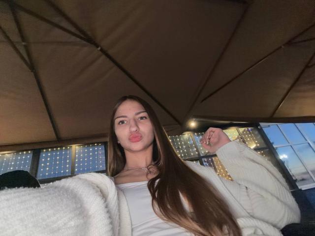 AdrianaNalley with long brown hair, puffy white cardigan, and white top, making a pouty face, under a tent at dusk, holding a ring on her right hand.