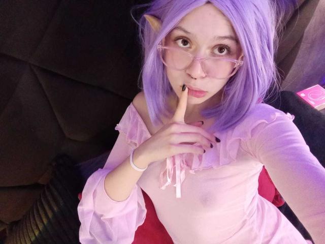 SophieBlake with purple hair, elf ears, pink glasses, and pink off-shoulder top, finger in mouth, showing small, visible nipples, sitting on dark couch.
