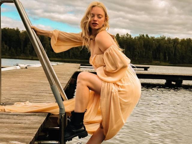 CrystalHope, blonde, off-shoulder peach dress, black boots, climbing metal ladder on wooden dock, water and forest in background, looking slightly annoyed, dress flowing in wind.