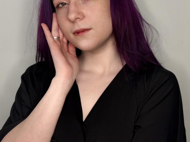 Margit Bossick, con i capelli viola, la pelle chiara e le labbra rosse, indossa una maglietta nera a V, con una mano sul viso e un anello d'argento, e sembra leggermente triste..