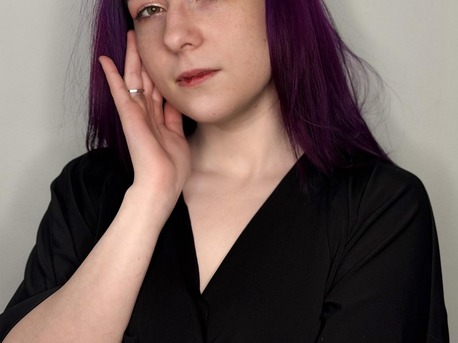 Margit Bossick, com cabelo roxo, pele clara e lábios vermelhos, vestindo uma blusa preta com decote em V, com a mão no rosto e um anel de prata, com uma expressão ligeiramente triste..